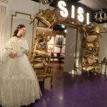 In einem Wachsfigurenkabinett sind die Figuren einer Frau in einem weißen Kleid und eines Mannes in Uniform zu sehen. Im Hintergrund befindet sich ein Bogen aus Stühlen und ein "SISI"-Schild.