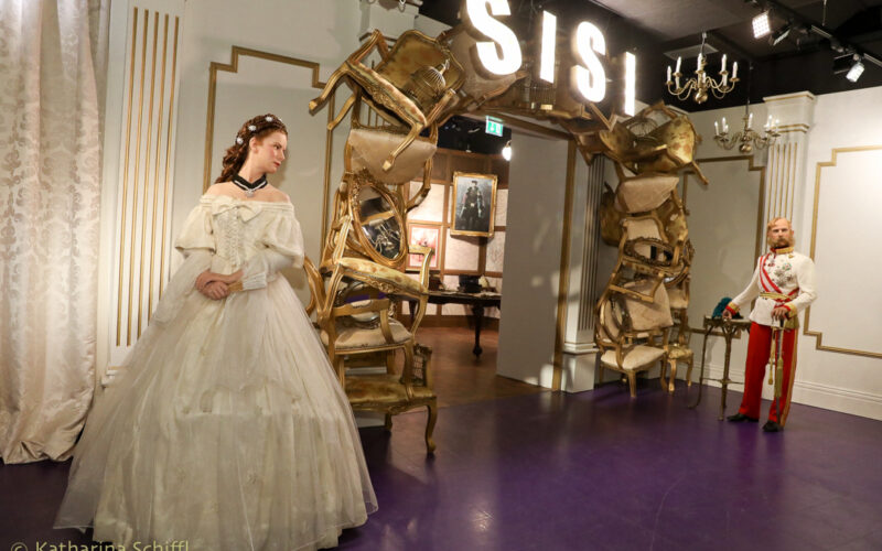 In einem Wachsfigurenkabinett sind die Figuren einer Frau in einem weißen Kleid und eines Mannes in Uniform zu sehen. Im Hintergrund befindet sich ein Bogen aus Stühlen und ein "SISI"-Schild.