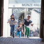 Drei Kinder, zwei davon in Ritterkostümen, laufen und lächeln auf einem Kopfsteinpflasterweg vor einem Gebäude mit der Aufschrift "MÜNZE HALL".