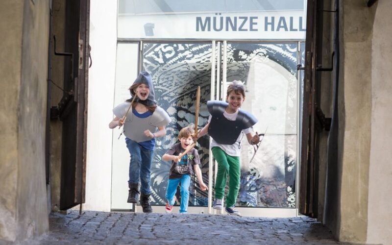 Drei Kinder, zwei davon in Ritterkostümen, laufen und lächeln auf einem Kopfsteinpflasterweg vor einem Gebäude mit der Aufschrift "MÜNZE HALL".