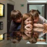 Drei Kinder untersuchen in einem Museum Gegenstände in einer Glasvitrine, wobei ihre Spiegelungen auf der Glasoberfläche sichtbar sind.
