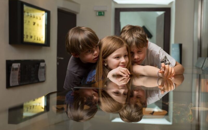 Drei Kinder untersuchen in einem Museum Gegenstände in einer Glasvitrine, wobei ihre Spiegelungen auf der Glasoberfläche sichtbar sind.