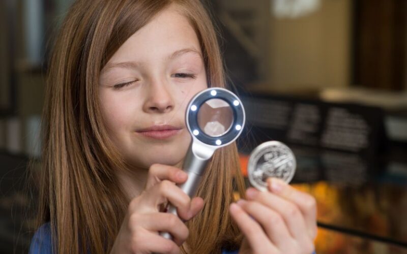 Ein Mädchen untersucht eine Münze mit einem Vergrößerungsglas, wobei es die Münze in der einen und die Lupe in der anderen Hand hält, vor einem unscharfen Hintergrund.