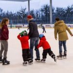Menschen verschiedenen Alters beim Schlittschuhlaufen in einer Eishalle, einige halten sich an den Händen, andere laufen unabhängig voneinander. Draußen sind Bäume und ein bewölkter Himmel durch Glasscheiben zu sehen.