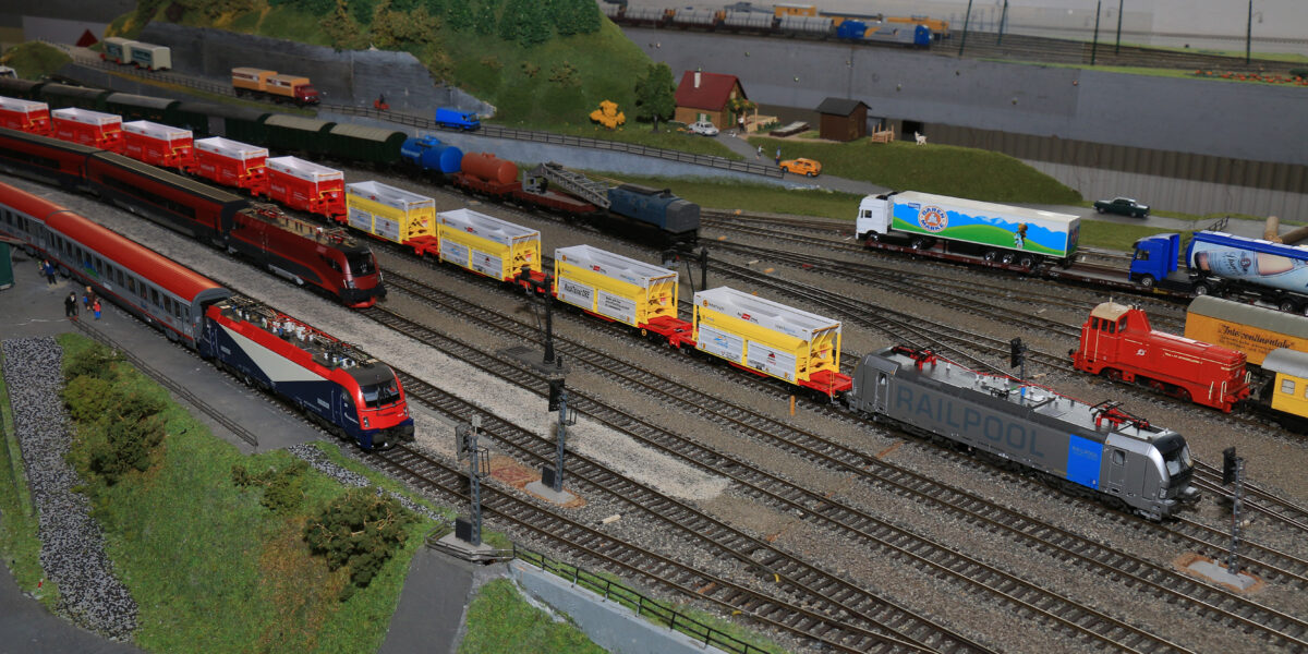 Modelleisenbahnanlage mit mehreren Gleisen, verschiedenen Modelleisenbahnen, Güterwagen und einer kleinen Grünanlage mit Bäumen und Miniaturgebäuden im Hintergrund.