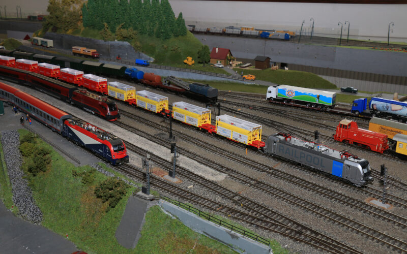Modelleisenbahnanlage mit mehreren Gleisen, verschiedenen Modelleisenbahnen, Güterwagen und einer kleinen Grünanlage mit Bäumen und Miniaturgebäuden im Hintergrund.