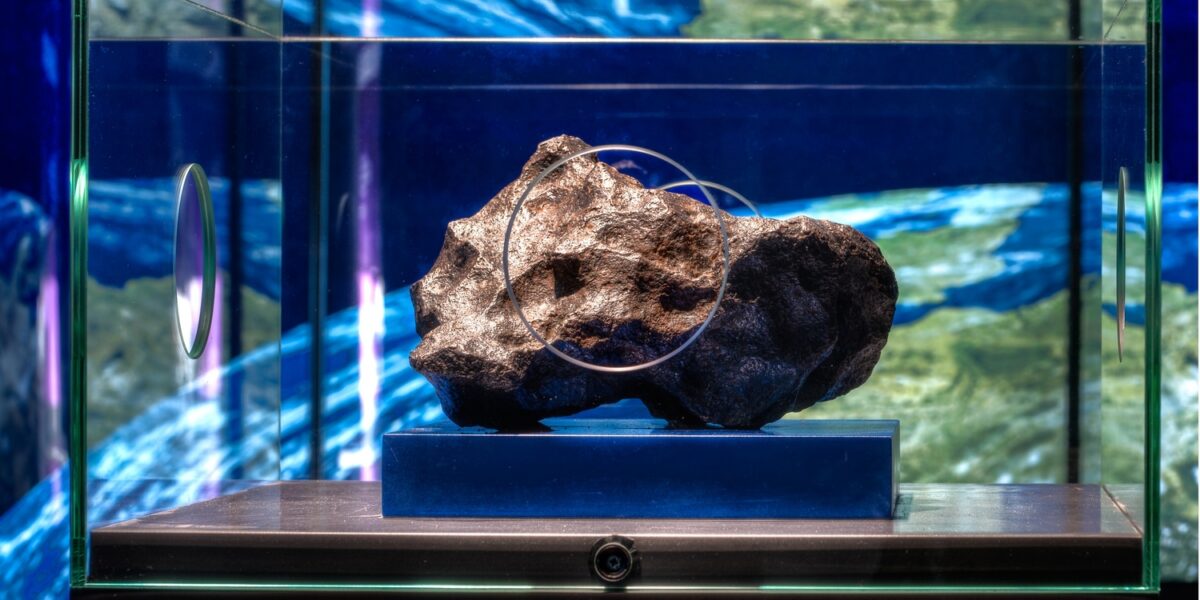Ein großer dunkler Meteorit wird auf einem blauen Ständer in einer Glasvitrine vor einem farbenfrohen, abstrakten Hintergrund ausgestellt.
