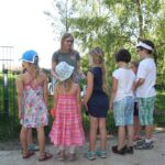 Eine Gruppe von Kindern hört an einem sonnigen Tag im Freien einer Frau im grünen Hemd an einem Metallzaun zu.