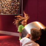 Ein junges Mädchen mit fliegenden Haaren steht vor einem großen Ventilator, der von einem anderen Kind bedient wird, in einem Raum mit roten Wänden und einer Kunstausstellung im Hintergrund.