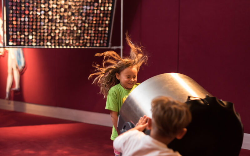 Ein junges Mädchen mit fliegenden Haaren steht vor einem großen Ventilator, der von einem anderen Kind bedient wird, in einem Raum mit roten Wänden und einer Kunstausstellung im Hintergrund.