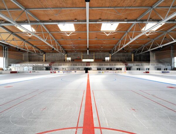 Leere Eishockeyhalle mit markierten Linien und Toren, umgeben von Sitzgelegenheiten und einer hohen Decke mit Sichtbalken.