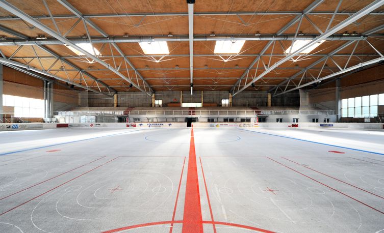 Leere Eishockeyhalle mit markierten Linien und Toren, umgeben von Sitzgelegenheiten und einer hohen Decke mit Sichtbalken.
