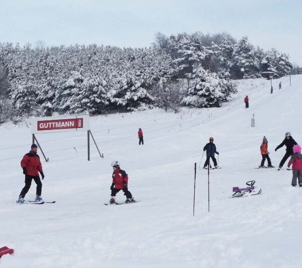 Menschen fahren Ski und Snowboard auf einer verschneiten, von Bäumen umgebenen Piste. Im Hintergrund ist ein rotes Schild mit der Aufschrift "GUTTMANN" zu sehen.