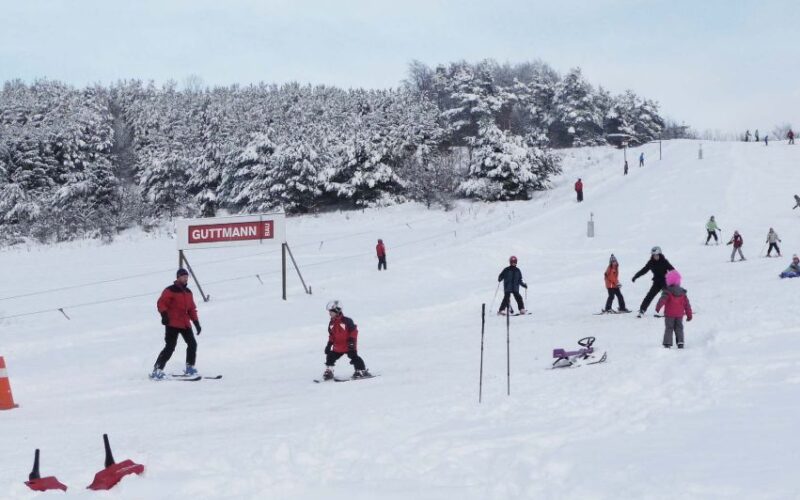Menschen fahren Ski und Snowboard auf einer verschneiten, von Bäumen umgebenen Piste. Im Hintergrund ist ein rotes Schild mit der Aufschrift "GUTTMANN" zu sehen.