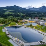 Luftaufnahme eines modernen Wellness-Resorts mit Außenpools, Lounge-Bereichen und einem natürlichen Teich in einem grünen, von Bergen umgebenen Tal in der Abenddämmerung.