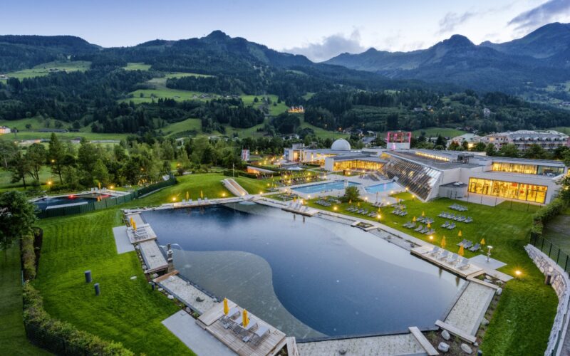 Luftaufnahme eines modernen Wellness-Resorts mit Außenpools, Lounge-Bereichen und einem natürlichen Teich in einem grünen, von Bergen umgebenen Tal in der Abenddämmerung.