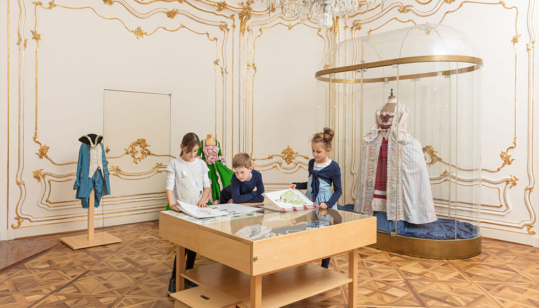 Drei Kinder sitzen an einem Zeichentisch in einem Museumsraum mit verschnörkelten Wänden, ausgestellten historischen Kostümen und einem Kronleuchter über dem Kopf.