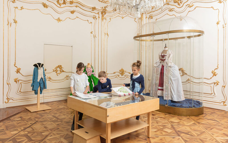 Drei Kinder sitzen an einem Zeichentisch in einem Museumsraum mit verschnörkelten Wänden, ausgestellten historischen Kostümen und einem Kronleuchter über dem Kopf.