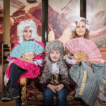 Drei Kinder in historischen Kostümen und Perücken sitzen auf einem Samtstuhl und halten dekorative Fächer in der Hand, während im Hintergrund ein Wandgemälde mit klassischen Figuren zu sehen ist.