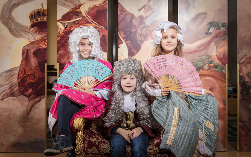Drei Kinder in historischen Kostümen und Perücken sitzen auf einem Samtstuhl und halten dekorative Fächer in der Hand, während im Hintergrund ein Wandgemälde mit klassischen Figuren zu sehen ist.