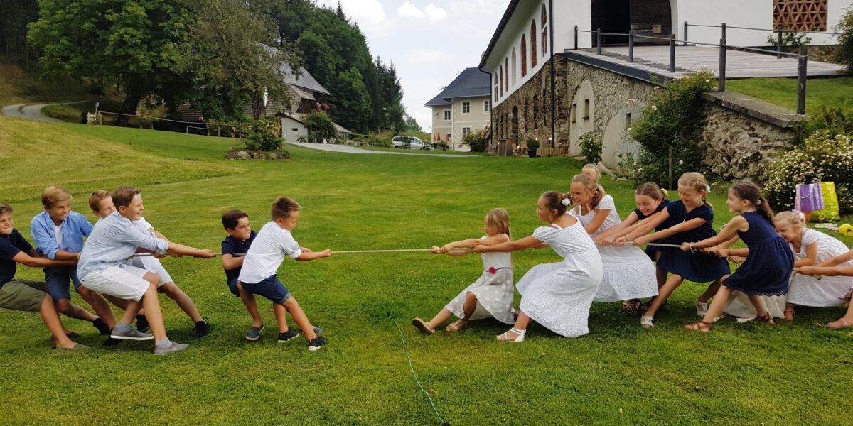 Eine Gruppe von Kindern, aufgeteilt in zwei Teams, spielt Tauziehen auf einer Wiese vor großen weißen Gebäuden.