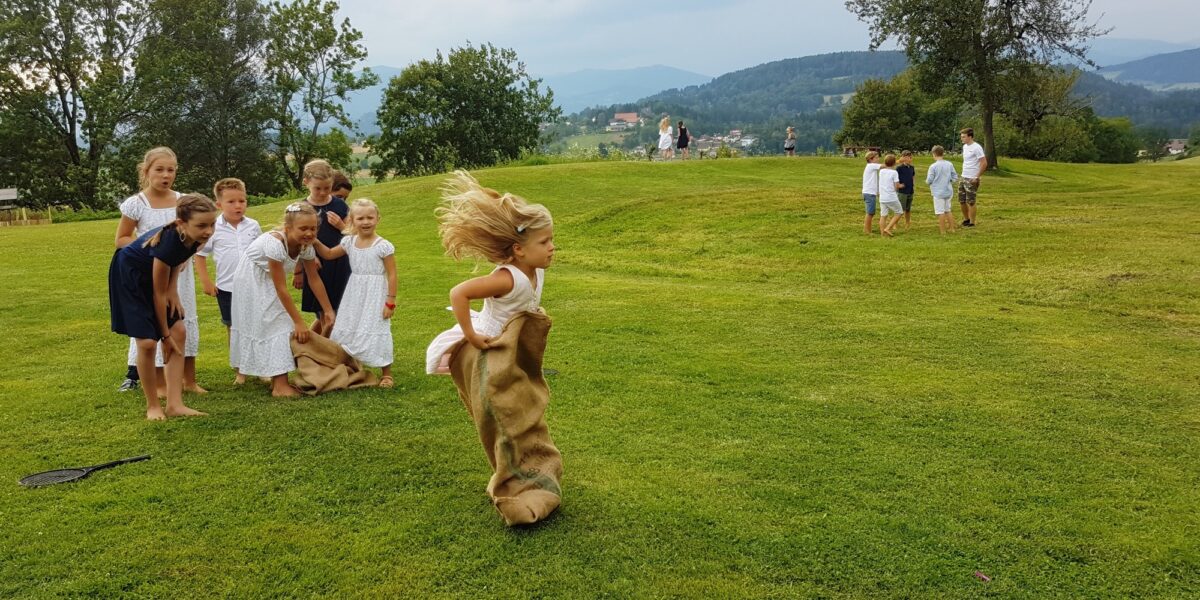 Kinder nehmen an einem Sackhüpfen auf einer Wiese teil, während sich im Hintergrund andere Kindergruppen versammeln. Das "Kinderwelt"-Logo ist in der unteren linken Ecke zu sehen.