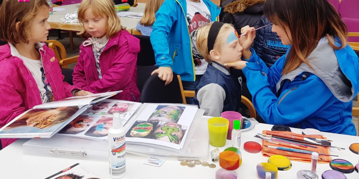 Eine Frau malt das Gesicht eines Jungen an einem mit Malutensilien gedeckten Tisch, während andere Kinder bei einer Veranstaltung mit dem Titel "Kinderwelt" zusehen und warten.