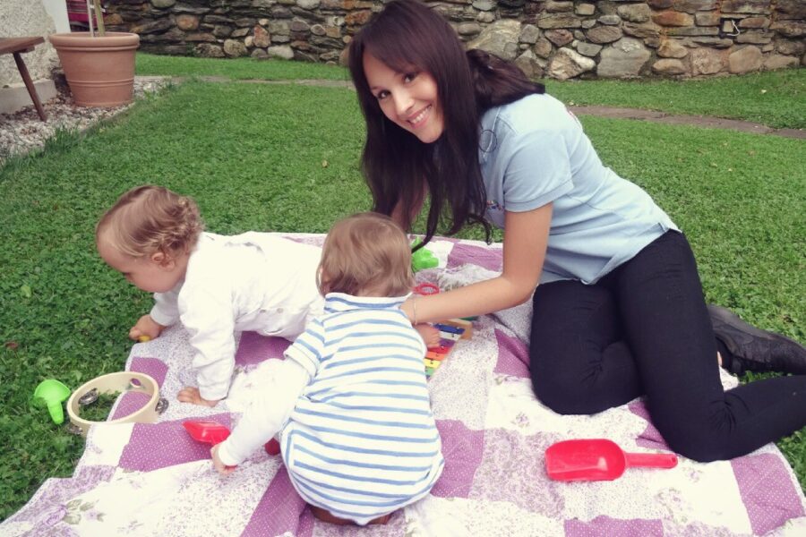 Eine Frau sitzt auf einer Decke im Freien mit zwei kleinen Kindern, die mit Spielzeug im Gras spielen.
