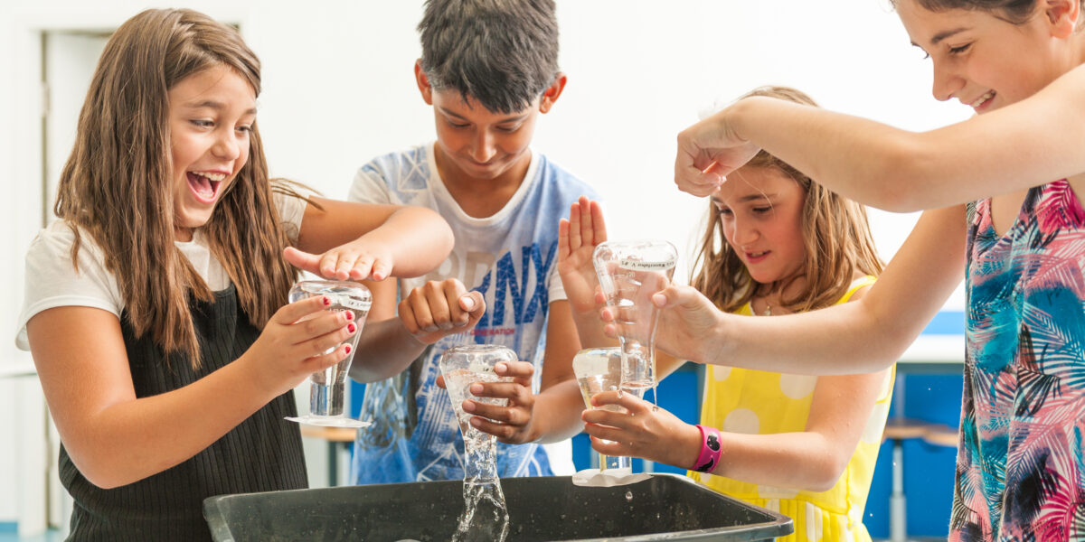 Vier Kinder führen ein praktisches wissenschaftliches Experiment mit Wasser und Plastikflaschen durch und stehen dabei in einem hell erleuchteten Klassenzimmer um eine schwarze Wanne herum.