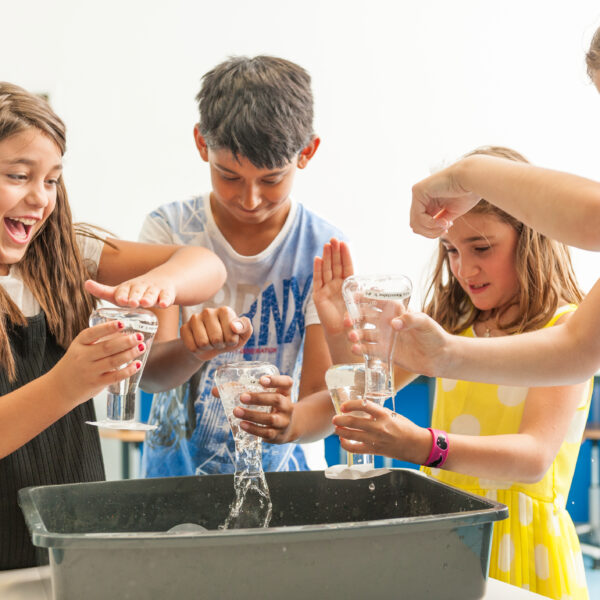 Vier Kinder führen ein praktisches wissenschaftliches Experiment mit Wasser und Plastikflaschen durch und stehen dabei in einem hell erleuchteten Klassenzimmer um eine schwarze Wanne herum.