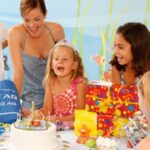 Eine Gruppe von Kindern und ein Erwachsener versammeln sich auf einer Geburtstagsfeier um einen Tisch mit einem Geburtstagskuchen, Geschenken und Dekorationen.