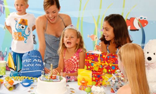 Eine Gruppe von Kindern und ein Erwachsener versammeln sich auf einer Geburtstagsfeier um einen Tisch mit einem Geburtstagskuchen, Geschenken und Dekorationen.