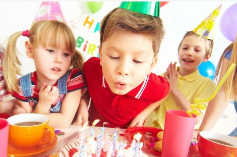 Drei Kinder mit Partyhüten sitzen an einem Tisch, während der Junge in der Mitte die Kerzen auf einer Geburtstagstorte auspustet.