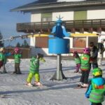 Eine Gruppe von Kindern in grünen Westen und Helmen nimmt an einem Skikurs auf einer verschneiten Piste in der Nähe eines Gebäudes teil, beaufsichtigt von Erwachsenen.