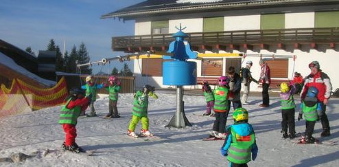 Eine Gruppe von Kindern in grünen Westen und Helmen nimmt an einem Skikurs auf einer verschneiten Piste in der Nähe eines Gebäudes teil, beaufsichtigt von Erwachsenen.
