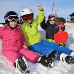 Drei Kinder in bunter Winterkleidung sitzen auf dem verschneiten Boden, lächeln und heben die Arme, eines hält einen roten Schneeschlitten.