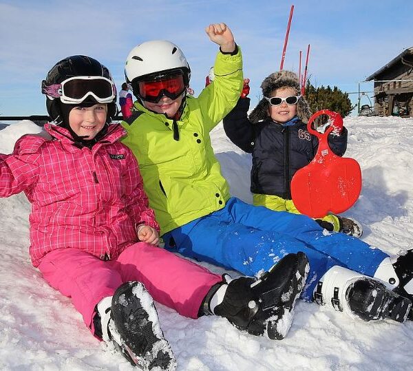 Drei Kinder in bunter Winterkleidung sitzen auf dem verschneiten Boden, lächeln und heben die Arme, eines hält einen roten Schneeschlitten.