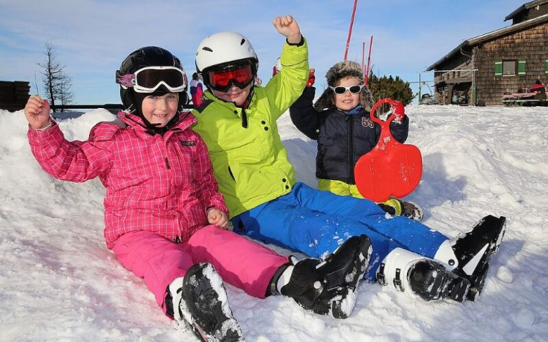 Drei Kinder in bunter Winterkleidung sitzen auf dem verschneiten Boden, lächeln und heben die Arme, eines hält einen roten Schneeschlitten.