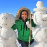 Eine Person in einer grünen Jacke steht draußen im Schnee zwischen zwei Schneemännern und lächelt in die Kamera.