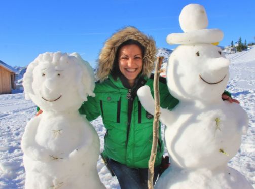 Eine Person in einer grünen Jacke steht draußen im Schnee zwischen zwei Schneemännern und lächelt in die Kamera.