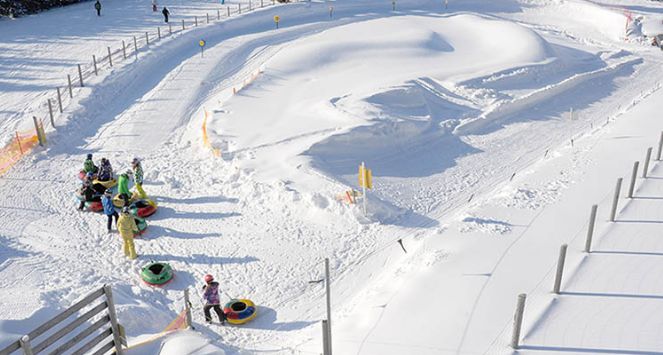 Kinder warten mit ihren Snowtubes am Fuße eines präparierten, verschneiten Tubing-Hügels, der von Zäunen und orangefarbenen Sicherheitsnetzen umgeben ist.