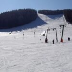 Skifahrer und Snowboarder benutzen einen Skilift auf einer verschneiten Skipiste mit einem bewaldeten Hügel im Hintergrund an einem klaren Tag.
