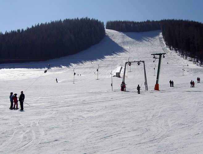 Skifahrer und Snowboarder benutzen einen Skilift auf einer verschneiten Skipiste mit einem bewaldeten Hügel im Hintergrund an einem klaren Tag.