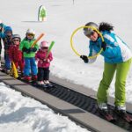 Ein Skilehrer führt vier kleine Kinder auf Skiern ein Förderband auf einer verschneiten Piste hinauf, in der Hand einen gelben Reifen und farbige Schaumstoffstöcke.