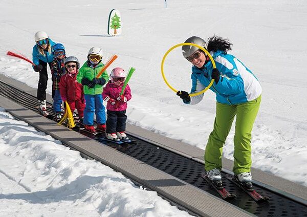 Ein Skilehrer führt vier kleine Kinder auf Skiern ein Förderband auf einer verschneiten Piste hinauf, in der Hand einen gelben Reifen und farbige Schaumstoffstöcke.