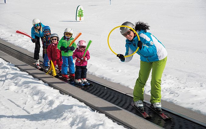 Ein Skilehrer führt vier kleine Kinder auf Skiern ein Förderband auf einer verschneiten Piste hinauf, in der Hand einen gelben Reifen und farbige Schaumstoffstöcke.