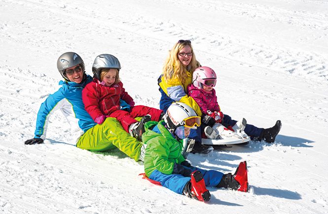 Vier Personen, zwei Erwachsene und zwei Kinder, sitzen in Winterkleidung und mit Helmen im Schnee und scheinen sich auf einer Skipiste auszuruhen oder zu posieren.