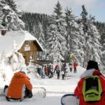 Menschen mit Snowboards sitzen und stehen in einem verschneiten Wald in der Nähe einer Hütte, vor der sich Gruppen versammelt haben; im Hintergrund sind schneebedeckte Bäume und ein klarer Himmel zu sehen.