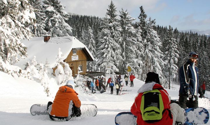 Menschen mit Snowboards sitzen und stehen in einem verschneiten Wald in der Nähe einer Hütte, vor der sich Gruppen versammelt haben; im Hintergrund sind schneebedeckte Bäume und ein klarer Himmel zu sehen.