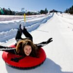 Eine Person lächelt, während sie an einem klaren Wintertag auf einem roten Schneeschlauch mit ausgestreckten Armen einen verschneiten Hügel hinunterrutscht.